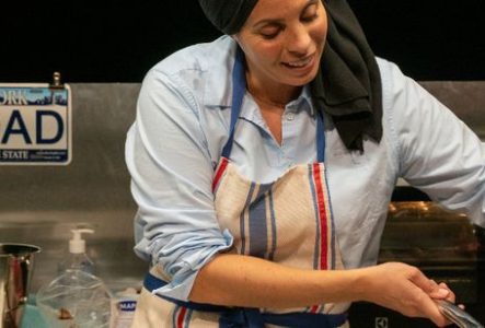 La photographie montre une personne derrière un plan de travail de cuisine installé sur une scène ou dans un espace de spectacle. La personne porte une chemise bleu clair, un tablier à rayures verticales bleu, blanc et rouge, ainsi qu’un tissu noir couvrant la tête et tombant sur une épaule.
Au premier plan, la personne utilise une spatule rouge au-dessus d’un récipient ou d’une préparation situé hors du cadre inférieur. Les mains sont occupées à une activité culinaire.
Derrière le plan de…