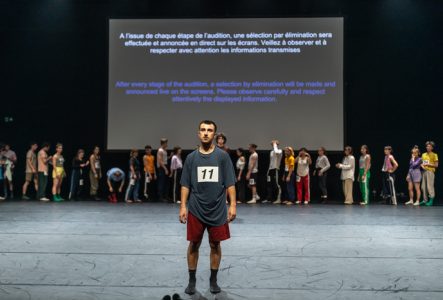 © Gregory Batardon Au premier plan, un danseur avec le numéro 11 sur son t-shirt, en attente, fait face au public. En arrière plan, une ligne de danseurs et de danseuses se préparant devant un écran sur lequel est projeté le texte suivant : A l'issue de chaque étape de l'audition, une sélection par élimination sera effectuée et annoncée en direct sur les écrans. Veillez à observer et à respecter avec attention les informations transmises.