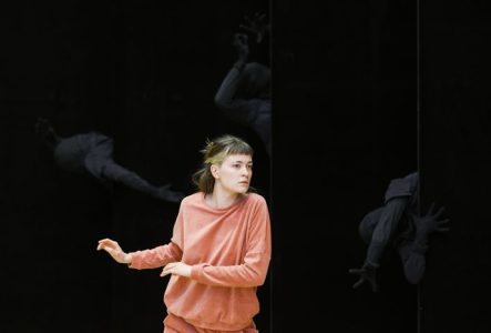 Une jeune femme porte un survêtement couleur saumon. À l’arrière-plan, la scène est sombre, mais on distingue plusieurs personnes vêtues de blanc.