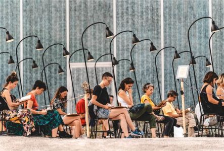 Un groupe de gens est assis sur des chaises sous des lampes - en extérieur - et chacun lit seul quelque chose devant lui.