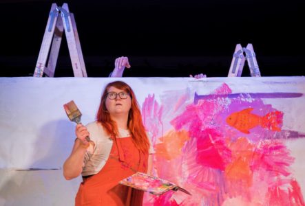 Une jeune femme avec un gros pinceau à la main devant une grande peinture