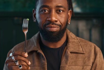 DR L'humoriste Christian Mukuna, nous regarde, il est habillé d'un veste couleur marron, tient verticalement dans sa main droite une fourchette à hauteur de son visage, avec planté au bout un unique petit pois.