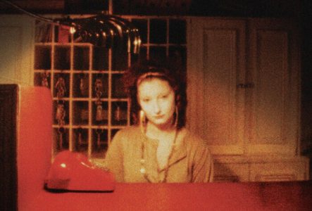 Affiche de film aux tons très sombres, dominée par des couleurs rouge foncé, brun et noir, avec une texture granuleuse des images de cinéma analogique. Au centre, une femme est assise derrière un comptoir d'hôtel rouge. Son visage est légèrement flou et éclairé par une lumière chaude qui crée une atmosphère nocturne et mystérieuse. Une lampe de bureau au design arrondi est posée à gauche, projetant une lumière tamisée. À l’arrière-plan, on distingue des étagères où des clés sont accrochées dans…