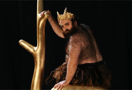 Un homme barbu portant une couronne dorée sur la tête et un pagne tient une (fausse) branche d'arbre dorée.