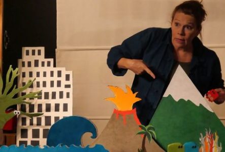 Une personne se tient derrière une maquette colorée représentant un paysage miniature. La personne porte une veste bleu foncé et tient dans une main un petit cube rouge. L’autre main est dirigée vers le décor, comme pour désigner un élément de la scène.
Au premier plan, plusieurs éléments découpés et peints sont disposés sur une table : à gauche, un arbre vert stylisé et un immeuble blanc à fenêtres noires ; au centre, une vague bleue et un volcan gris en éruption, dont le sommet laisse jaillir…