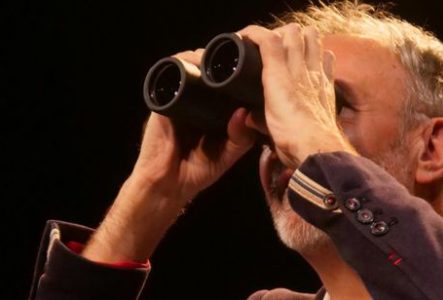 Gros plan d’une personne sur une scène sombre, éclairée par une lumière chaude. La personne porte une veste foncée avec des boutons visibles aux poignets et tient une paire de jumelles noires devant les yeux à l’aide de ses deux mains.
Le cadrage est serré sur le haut du corps et le visage est en grande partie masqué par les jumelles. On distingue des cheveux courts et une barbe grisonnante. Les bras sont relevés pour maintenir les jumelles en position d’observation.
L’arrière-plan est…
