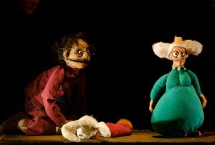 La photographie montre une scène de théâtre de marionnettes éclairée par une lumière chaude. Au centre, sur un plateau en bois, se trouvent deux marionnettes de taille moyenne.
À gauche, une marionnette porte un vêtement rouge foncé à rayures. La marionnette est agenouillée au sol et porte de grandes lunettes rondes. À droite, une seconde marionnette est vêtue d’un costume vert turquoise volumineux et possède de très grandes oreilles blanches qui s’étendent de chaque côté de la tête.
Entre les…