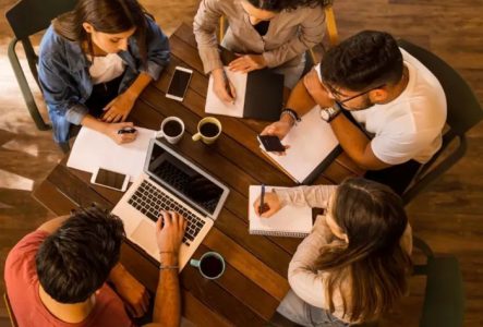 Vue aérienne en plongée de cinq participants à un atelier d'écriture, réunis autour d'une table carrée en bois chaleureux. Dans une ambiance de travail collaborative et conviviale, trois femmes et un homme écrivent à la main avec concentration dans des cahiers ou sur des feuilles blanches, tandis qu'un homme utilise un ordinateur portable. Quelques tasses de café et des téléphones portables sont disposés sur la table, illustrant un moment de création littéraire partagé.