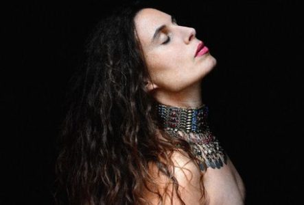 Portrait en gros plan d’une personne photographiée de profil sur un fond entièrement noir. La tête est légèrement inclinée vers l’arrière et les yeux sont fermés. Les cheveux longs et ondulés tombent sur les épaules.
La personne porte un imposant collier métallique couvrant largement le cou et le haut de la poitrine. Ce bijou est composé de multiples rangées d’éléments argentés ornés de petites pierres colorées, avec plusieurs pendentifs suspendus à l’avant.
L’éclairage met en valeur le visage,…