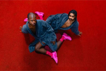 Deux hommes en peignoir bleu avec des baskets roses regardent la caméra. Ils sont allongés sur un tapis rouge.