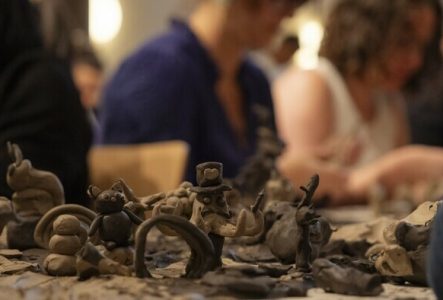 Au premier plan, on voit des animaux ou d'autres éléments modelés en terre. A l'arrière plan, on voit des personnes floues en train de travailler la terre.