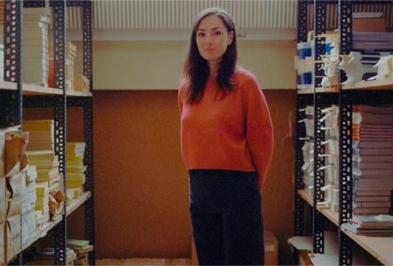Une femme en pull rouge se tient entre deux étagères recouvertes de manuscrits de livres.