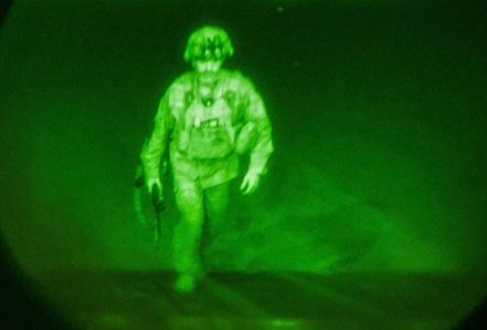 Photographie en vue nocturne en tons de vert montrant un soldat américain marchant avec un fusil à la main en direction de la camera.