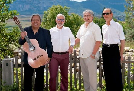 Quatre hommes souriants debout dont un avec une guitare en main