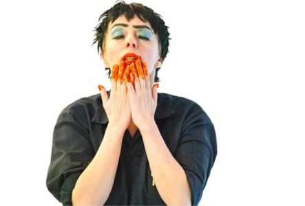 Une femme met ses mains à son visage, elles sont maculées de peinture orange. La femme est sur un fond blanc avec une blouse noire.