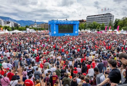 DR Une foule dense de milliers de personnes, majoritairement vêtues de rouge, est rassemblée en plein air devant un grand écran bleu marqué « Fan Zone Genève 2024 ». L’ambiance est festive. Des tentes blanches bordent les côtés, et des montagnes sont visibles en arrière-plan sous un ciel nuageux. On aperçoit aussi des drapeaux, des bâtiments et une église au loin.