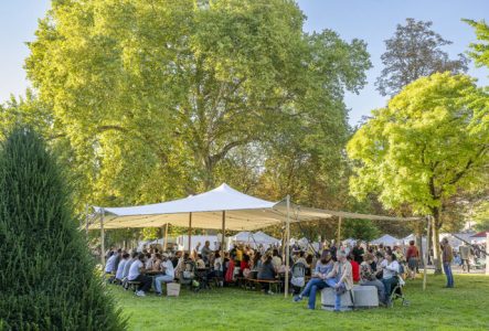 L'image montre une grande tente blanche, installée sur une vaste pelouse verte. Sous cette tente ouverte, de nombreuses personnes sont assises sur des bancs autour de longues tables en bois, partageant un repas.