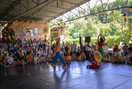 Quatre danseuses se tiennent sur une scène au premier plan. En second plan, on distingue le public assis, composé de nombreuses personnes. La scène se situe dans un hangar ouvert, rappelant une ancienne structure industrielle. On observe que le lieu a été transformé à des fins socio-culturelles, avec la présence de petites constructions.