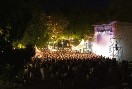 DR On voit une foule rassemblée devant une scène illuminée en pleine nuit, baignée de lumières vives. Des arbres entourent la scène, ajoutant une touche naturelle à l’ambiance festive.