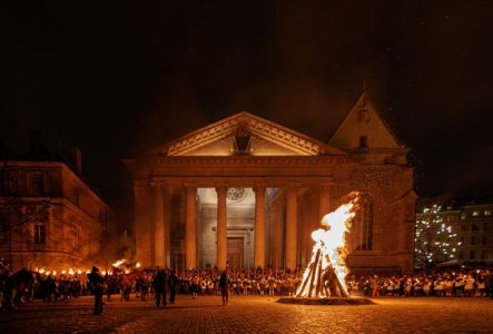 Un grand feu de joie brûle sur sur le parvis de la Cathédrale Saint-Pierre. Une foule nombreuse, dont beaucoup de porteurs de flambeaux et de torches, entoure le brasier dans la nuit. On distingue aussi le Héraut et son cheval au premier plan. La scène dégage une ambiance très solennelle et festive, typique d'une commémoration nocturne comme celle de l'Escalade à Genève.