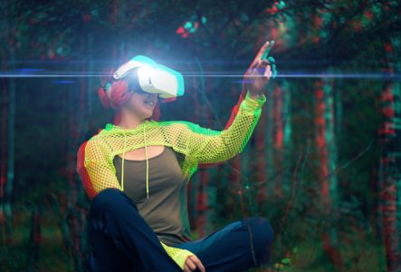 123rf Une femme assise dans l'herbe, habits futuristes ou sport technique, un casque VR sur les yeux, lève le doigt comme pour toucher quelque chose devant elle. C'est dans la forêt, l'image est glitchée, comme décalée en rouge et bleu.
