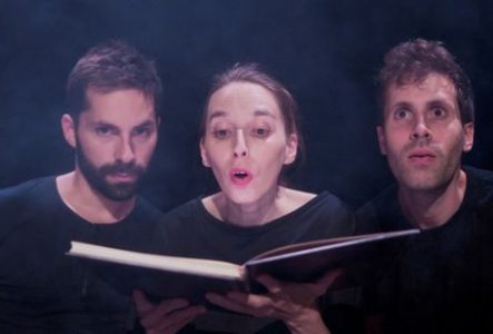 Photographie prise dans un environnement sombre, probablement une scène de théâtre. Trois personnes sont réunies au premier plan, côte à côte, devant un grand livre ouvert tenu au centre de l’image.
Les trois personnes portent des vêtements foncés. La personne au centre tient le livre ouvert avec les deux mains, tandis que les deux autres se penchent également vers l’ouvrage. Le livre est orienté vers les personnes et son contenu n’est pas visible.
L’éclairage est concentré sur les visages, les…