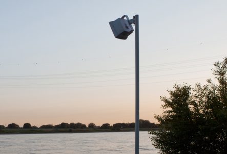Sur cette image on voit un haut-parleur fixé en haut d’un mât métallique, installé au bord d’un fleuve.