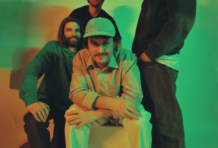 II (c) Midle Photo promotionnelle de l’événement Jazztronica #3. Quatre musiciens posent dans un studio éclairé de lumières vertes et orangées. Un membre est assis au centre, casquette claire et chemise beige, les trois autres se tiennent debout derrière lui, souriants, en tenues décontractées.