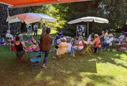 Paola Emery Tables rondes et chaises avec des parasols disposées dans le jardin. Des gens écoutant des orateurs