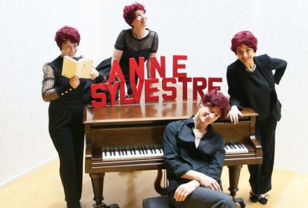 Au centre, un piano à queue, sur lequel est posé de grosses lettres rouges formant le nom d'Anne Sylvestre. Autour, 4 personnes avec des perruques imitant les cheveux courts et rouges d'Anne Sylvestre.
