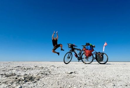 Un homme saute de joie dans un désert blanc, à côté de son vélo. Le ciel est d'un bleu intense en arrière-fond.