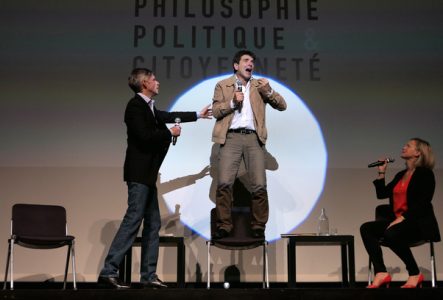 Un intervenant très engagé debout sur une chaise face à l'audience parle dans un micro et deux de ses collègues essaient de le calmer.