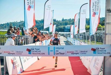 DR Sur l'image, on voit l'arrivée du Tour Genève Triathlon avec un coureur arrivant seul. De chaque côté, une foule l'encourage et des drapeaux portant le nom de l'événement sont visibles. Un tapis rouge est déposé sur le sol à l'arrivée. Le ciel est bleu, et en arrière-plan, on aperçoit des arbres verts.