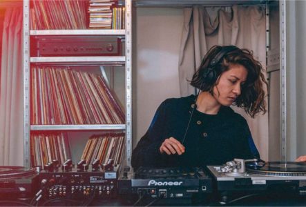 Une femme mix sur des platines de DJ dos à une étagère recouverte de CDs et vinyles.