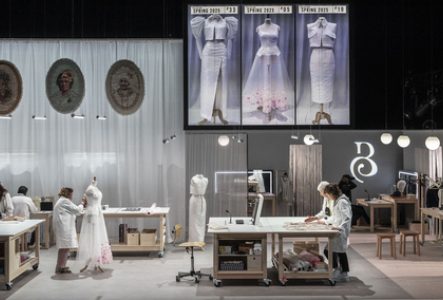 Sur cette image, nous pouvons voir plusieurs personnes travaillant dans un atelier de couture sur différents modèles et à différents endroits de la pièce. Il y a également en second plan sur un écran l'image de 3 robes.