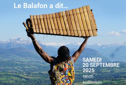 Educafon / Ecolint Un homme noir, habillé avec une toge multicolor qui brandit un grand balafon au dessus de sa tête en regardant vers le ciel, face à une plaine des montagnes et le ciel.