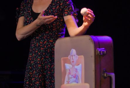 Jeune femme debout à côté d'une valise où est projetée l'image d'une femme couchée. La femme couchée est un personnage en pâte  à modeler