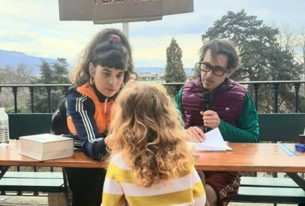 Cie Sale Bête production Deux adultes assis derrière une table regardent une enfant, dos à nous, de l'autre côté de la table. L'homme a un micro. Derrière eux, écriteau "Bureau des plaintes"