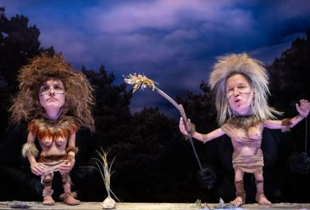 Deux personnages préhistoriques féminins en marionnettes Kokoschka font face au public. L'une d'entre-elles tient dans sa main un bâton en feu.