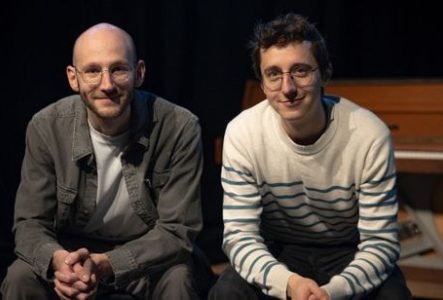 Photographie prise sur une scène de théâtre ou dans un espace de spectacle faiblement éclairé. Deux personnes sont assises côte à côte au premier plan, face à l’objectif.
La personne située à gauche porte des lunettes rondes, un t-shirt clair sous une veste grise et un pantalon assorti. La tête est rasée. Les mains sont jointes devant les genoux.
La personne située à droite porte également des lunettes rondes, un pull blanc à rayures horizontales bleues et un pantalon foncé. Les avant-bras…