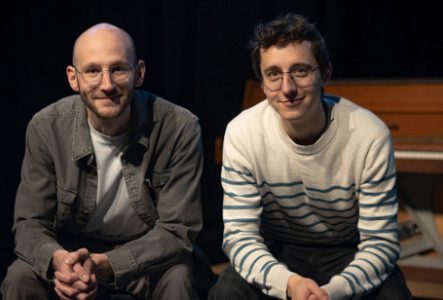 Photographie prise sur une scène de théâtre ou dans un espace de spectacle faiblement éclairé. Deux personnes sont assises côte à côte au premier plan, face à l’objectif.
La personne située à gauche porte des lunettes rondes, un t-shirt clair sous une veste grise et un pantalon assorti. La tête est rasée. Les mains sont jointes devant les genoux.
La personne située à droite porte également des lunettes rondes, un pull blanc à rayures horizontales bleues et un pantalon foncé. Les avant-bras…