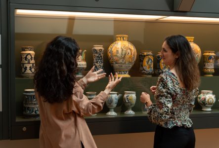 Une médiatrice donne des informations à une visiteuse sur une vitrine présentant des pots de pharmacie.