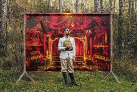 Un homme dans une forêt se tient devant une toile représentant une salle de théâtre dans les teintes rouges / or.