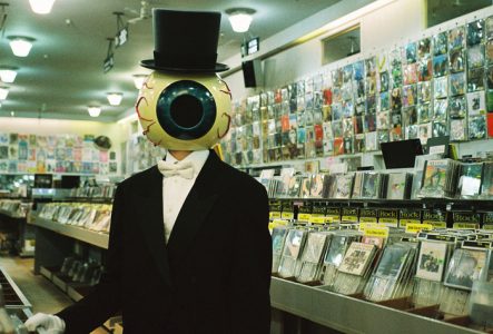 Sur cette image, on voit une personne avec un gros œil à la place de la tête. Elle porte un costume noir et fixe la caméra, dans un magasin de disques