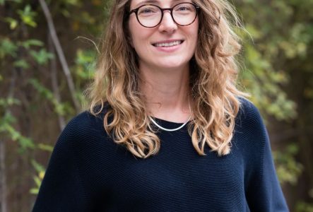 Portrait de l'intervenante jusqu'à la taille. Cheveux longs plutôt bouclés clair. Elle a un pull bleu foncé les mains dans les poches, des lunettes rondes et elle sourit.