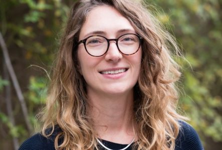 Portrait de l'intervenante jusqu'à la taille. Cheveux longs plutôt bouclés clair. Elle a un pull bleu foncé les mains dans les poches, des lunettes rondes et elle sourit.