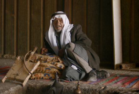 vieil homme, tenue traditionnelle, keffieh blanc (Foulard du Moyen-Orient), regard pensif, intérieur sombre, tapis coloré, coussins à motifs, instrument à cordes, artisanat, ambiance rustique, vêtements amples, bâton, lumière naturelle, regard
