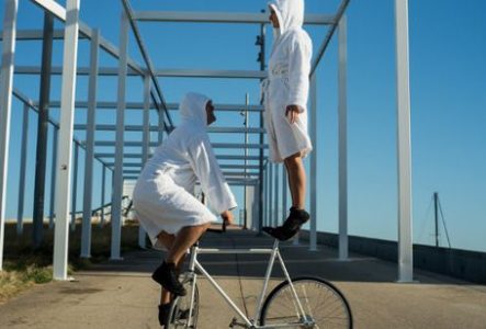 Photographie prise en extérieur sous un ciel bleu dégagé. Au centre de l’image, deux personnes vêtues de peignoirs blancs à capuche et de chaussures noires interagissent avec un vélo blanc.
Une personne est accroupie sur les pédales du vélo, maintenant l’équilibre. L’autre personne se tient debout sur le guidon et semble légèrement surélevée au-dessus du vélo. L’ensemble évoque une figure d’équilibre ou une acrobatie.
La scène se déroule sur une allée en béton bordée d’herbe. Autour des…