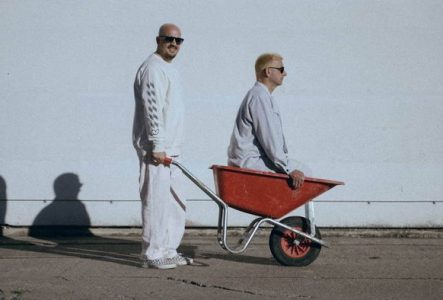 Photo des deux membres du groupe dont l'un pousse une bouette avec l'autre à l'intérieur. Ils sont habillés en blanc et portent des lunettes de soleil
