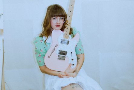 Moictani est montrée accroupie sur ses talons devant un tissu blanc avec sa guitare éléctrique rose claire dans les bras. Son visage est posé sur le haut de l´instrument et elle regarde la camera en face. Elle est habillée d´une jupe blanche, d´un chemisier vert clair avec un motif de fleurs rouges et de chaussures à talons épais rouges cerise. Elle porte de grandes lunettes de vue rondes et a les cheveux châtains dorés à la longueur de ses épaules avec une frange recouvrant son font.